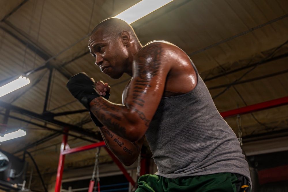 A muscular, bald man is training in a boxing gym, captured from a low angle. He is in a fighting stance with black hand wraps on his fists, wearing a gray sleeveless shirt and green shorts. His body is visibly sweating, and he has extensive tattoos on both arms.