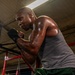 A muscular, bald man is training in a boxing gym, captured from a low angle. He is in a fighting stance with black hand wraps on his fists, wearing a gray sleeveless shirt and green shorts. His body is visibly sweating, and he has extensive tattoos on both arms.