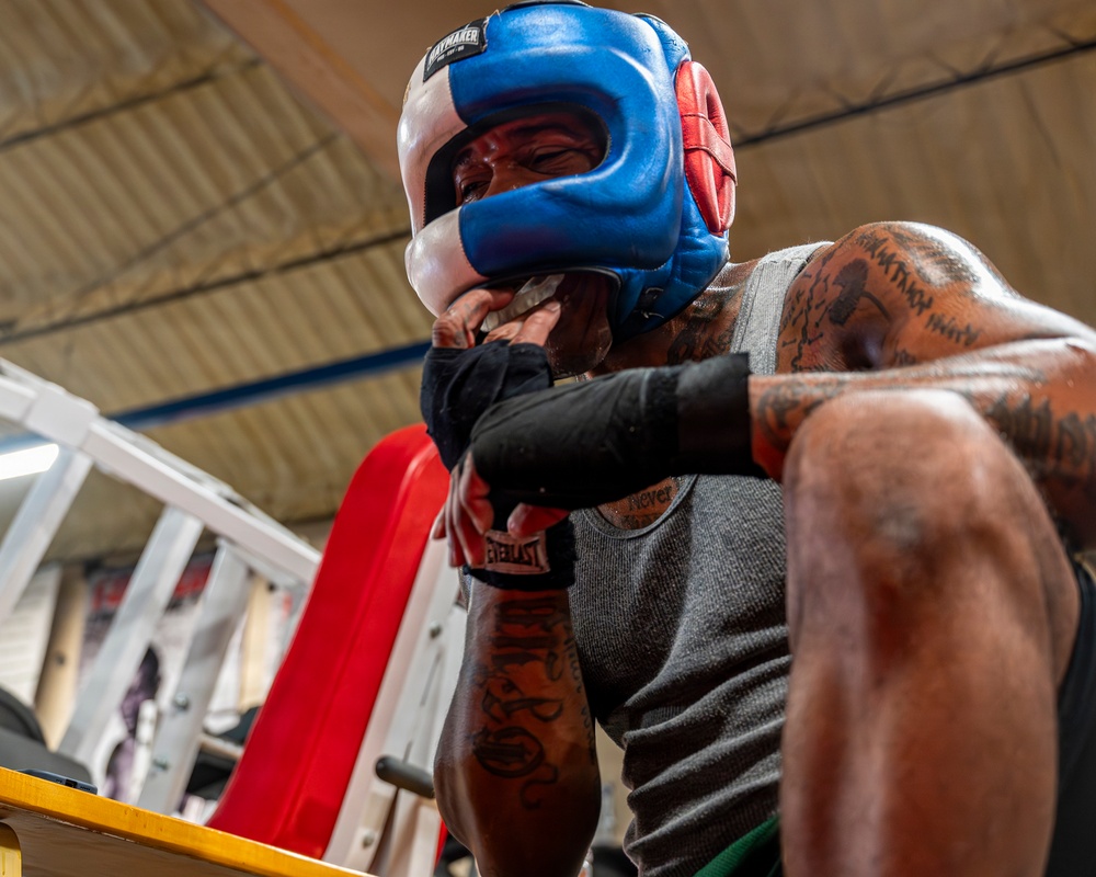 A heavily tattooed boxer is photographed from a low angle inside a boxing gym, wearing a blue and white headgear branded 'Haymaker' and black Everlast hand wraps in a defensive guard position. The athlete is leaning against what appears to be the ropes or corner of a boxing ring, wearing a gray sleeveless shirt. The image captures an intense training moment with dramatic lighting and perspective.