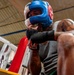 A heavily tattooed boxer is photographed from a low angle inside a boxing gym, wearing a blue and white headgear branded 'Haymaker' and black Everlast hand wraps in a defensive guard position. The athlete is leaning against what appears to be the ropes or corner of a boxing ring, wearing a gray sleeveless shirt. The image captures an intense training moment with dramatic lighting and perspective.