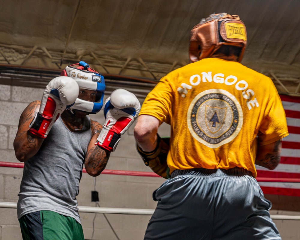 Two boxers are sparring in a boxing ring, both wearing headgear and boxing gloves. The boxer facing away wears a yellow t-shirt with 'MONGOOSE' printed on the back along with what appears to be a military unit insignia (25th Infantry Division). An American flag is visible in the background, and the setting appears to be an indoor boxing gym.