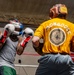 Two boxers are sparring in a boxing ring, both wearing headgear and boxing gloves. The boxer facing away wears a yellow t-shirt with 'MONGOOSE' printed on the back along with what appears to be a military unit insignia (25th Infantry Division). An American flag is visible in the background, and the setting appears to be an indoor boxing gym.
