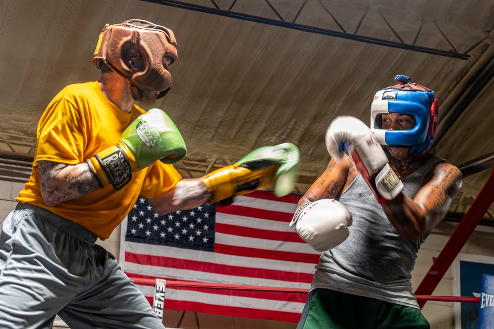 Two boxers are sparring in a boxing ring during a training session. The left boxer wears a yellow shirt with a brown leather headguard and green/yellow gloves, while the right boxer wears a gray tank top with a blue/red headguard and white gloves. An American flag is prominently displayed on the wall in the background.