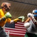 Two boxers are sparring in a boxing ring during a training session. The left boxer wears a yellow shirt with a brown leather headguard and green/yellow gloves, while the right boxer wears a gray tank top with a blue/red headguard and white gloves. An American flag is prominently displayed on the wall in the background.