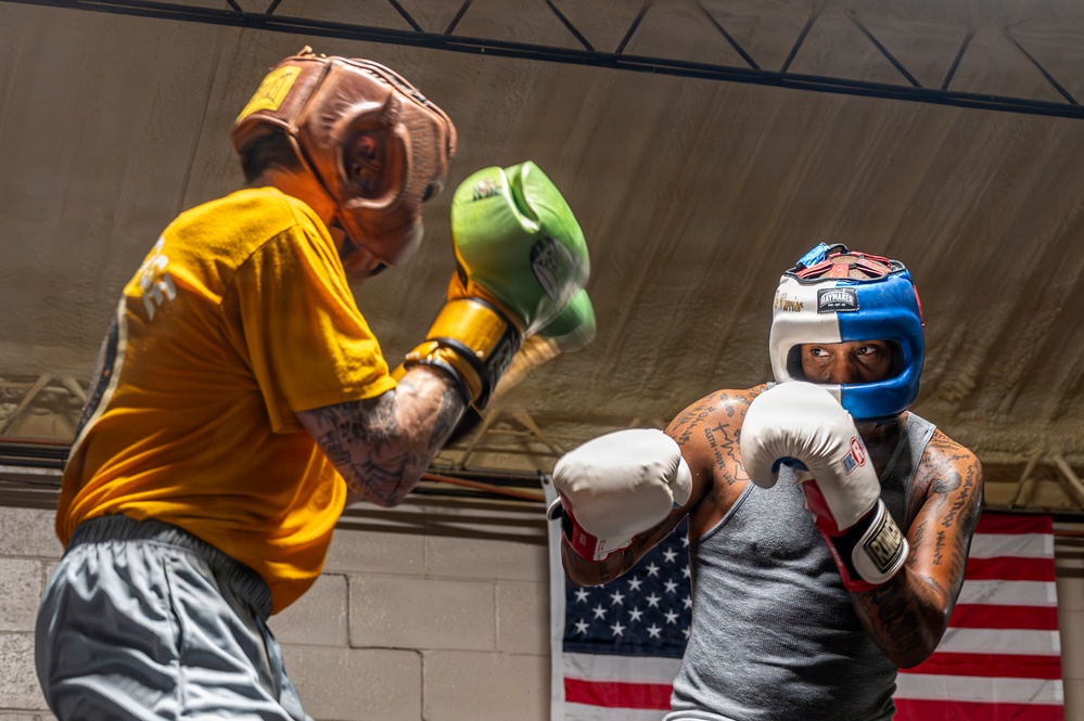 Two boxers are sparring in what appears to be a boxing gym or training facility. The boxer on the right, wearing a blue and white headgear and white gloves, is in a defensive stance while the boxer on the left, wearing a brown headgear and green gloves with a yellow shirt, is throwing a punch. An American flag is visible hanging on the wall in the background.