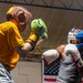 Two boxers are sparring in what appears to be a boxing gym or training facility. The boxer on the right, wearing a blue and white headgear and white gloves, is in a defensive stance while the boxer on the left, wearing a brown headgear and green gloves with a yellow shirt, is throwing a punch. An American flag is visible hanging on the wall in the background.