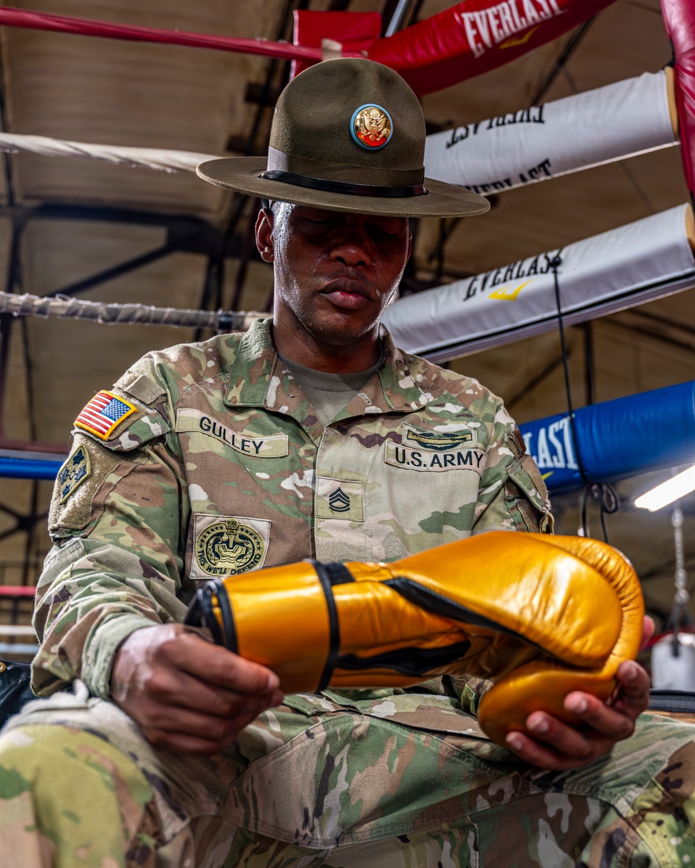 A U.S. Army Sergeant named Gulley, wearing an OCP (Operational Camouflage Pattern) uniform and a Drill Sergeant campaign hat, sits inside a boxing ring examining a pair of gold/yellow boxing gloves. His uniform displays a U.S. Army tape, name tape, Staff Sergeant rank chevrons, an American flag patch, and what appears to be a unit patch. The setting is a boxing gym with Everlast-branded ropes and equipment visible in the background.