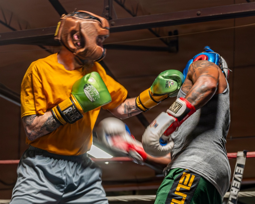 Two boxers are engaged in an active sparring session inside a boxing ring. The left boxer wears a yellow shirt, brown leather headgear, and green WBC-branded gloves, while the right boxer wears a gray tank top, blue headgear, and white/red/blue Everlast gloves. Motion blur on one glove indicates a punch in progress, capturing the dynamic action of the exchange.