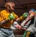 Two boxers are engaged in an active sparring session inside a boxing ring. The left boxer wears a yellow shirt, brown leather headgear, and green WBC-branded gloves, while the right boxer wears a gray tank top, blue headgear, and white/red/blue Everlast gloves. Motion blur on one glove indicates a punch in progress, capturing the dynamic action of the exchange.