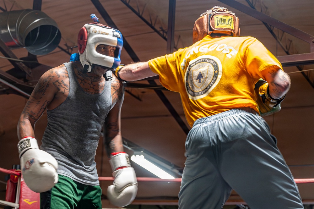 Two boxers are sparring inside a boxing gym ring. The fighter on the left wears a gray tank top, green shorts, white boxing gloves, and a white/red headgear, while the fighter on the right wears a yellow 'Mongoose' t-shirt, gray pants, gold/brown Everlast headgear, and dark gloves. The right fighter is throwing a punch toward the left fighter's head.