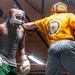 Two boxers are sparring inside a boxing gym ring. The fighter on the left wears a gray tank top, green shorts, white boxing gloves, and a white/red headgear, while the fighter on the right wears a yellow 'Mongoose' t-shirt, gray pants, gold/brown Everlast headgear, and dark gloves. The right fighter is throwing a punch toward the left fighter's head.
