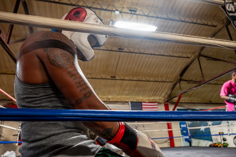 A boxer wearing a white and red headguard and a gray sleeveless shirt rests against the ropes in the corner of a boxing ring. The subject has visible tattoos on both arms and is wearing hand wraps. The setting appears to be an indoor boxing gym with an American flag visible in the background.
