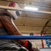 A boxer wearing a white and red headguard and a gray sleeveless shirt rests against the ropes in the corner of a boxing ring. The subject has visible tattoos on both arms and is wearing hand wraps. The setting appears to be an indoor boxing gym with an American flag visible in the background.