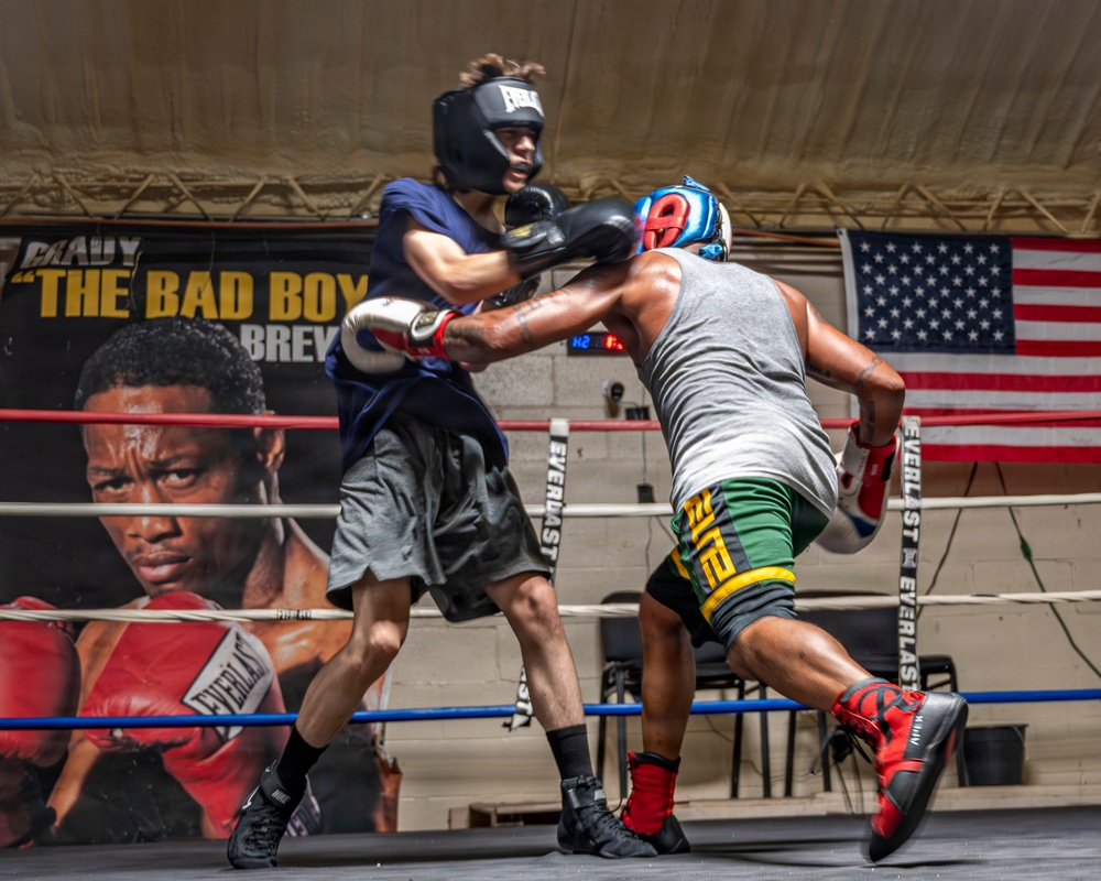 Two boxers are sparring in a boxing ring during what appears to be a training session. One boxer wearing a navy blue shirt and black headgear is exchanging punches with an opponent in a gray tank top and colorful green/yellow shorts with a blue and red headgear. The session takes place in a gym decorated with an American flag and a promotional banner reading 'The Bad Boy' with a fighter's portrait.