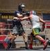 Two boxers are sparring in a boxing ring during what appears to be a training session. One boxer wearing a navy blue shirt and black headgear is exchanging punches with an opponent in a gray tank top and colorful green/yellow shorts with a blue and red headgear. The session takes place in a gym decorated with an American flag and a promotional banner reading 'The Bad Boy' with a fighter's portrait.