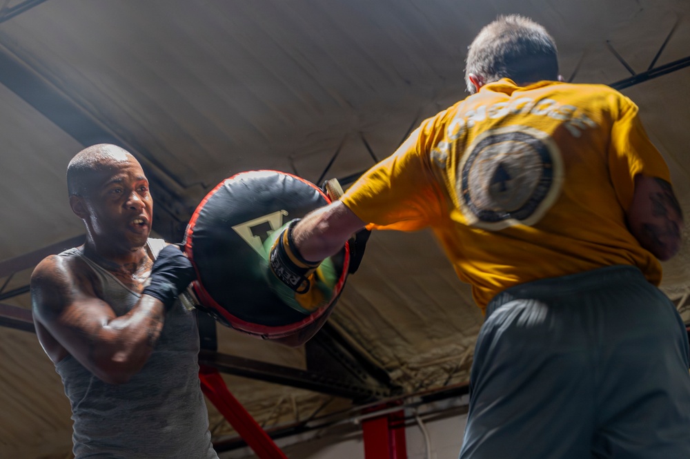 Two men are training in a boxing gym, with one person wearing a yellow t-shirt holding a large circular punch mitt while the other, wearing a gray tank top, delivers a punch. The session appears to be a pad-work training drill inside what looks like a boxing ring. The image is captured from a low angle, giving a dynamic and intense perspective to the action.