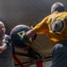 Two men are training in a boxing gym, with one person wearing a yellow t-shirt holding a large circular punch mitt while the other, wearing a gray tank top, delivers a punch. The session appears to be a pad-work training drill inside what looks like a boxing ring. The image is captured from a low angle, giving a dynamic and intense perspective to the action.