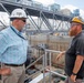 Two workers wearing hard hats are engaged in conversation at an elevated position overlooking a large industrial or civil engineering construction site, likely a dry dock, lock, or dam facility. The man on the left wears a white hard hat marked '700' and a plaid shirt, while the man on the right wears a decorated yellow hard hat marked '74' and a dark shirt. The background reveals extensive steel framework, scaffolding, concrete walls, and heavy construction equipment.