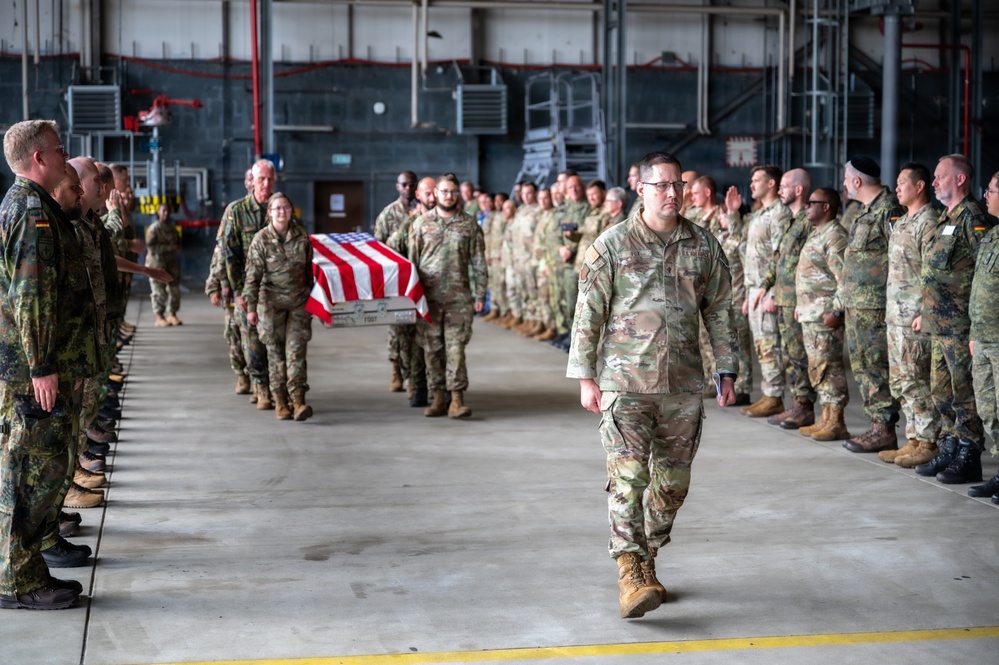 A solemn military dignified transfer or memorial ceremony is taking place inside a large hangar facility. A flag-draped casket is being carried by pallbearers down a corridor formed by two rows of standing military personnel, including both U.S. Army soldiers in OCP (Operational Camouflage Pattern) uniforms and German Bundeswehr soldiers in their distinctive flecktarn camouflage. A lead soldier walks ahead of the casket procession while personnel on both sides render salutes.
