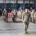 A solemn military dignified transfer or memorial ceremony is taking place inside a large hangar facility. A flag-draped casket is being carried by pallbearers down a corridor formed by two rows of standing military personnel, including both U.S. Army soldiers in OCP (Operational Camouflage Pattern) uniforms and German Bundeswehr soldiers in their distinctive flecktarn camouflage. A lead soldier walks ahead of the casket procession while personnel on both sides render salutes.