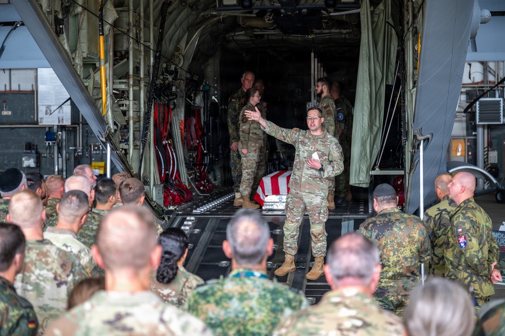 A military briefing or ceremony is taking place at the rear cargo ramp of a large military transport aircraft, likely a C-130 Hercules. A soldier in OCP (Operational Camouflage Pattern) uniform is addressing a large crowd of multinational military personnel. A flag-draped case or casket is visible in the background on the aircraft ramp, suggesting this may be a dignified transfer or memorial ceremony.