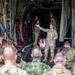 A military briefing or ceremony is taking place at the rear cargo ramp of a large military transport aircraft, likely a C-130 Hercules. A soldier in OCP (Operational Camouflage Pattern) uniform is addressing a large crowd of multinational military personnel. A flag-draped case or casket is visible in the background on the aircraft ramp, suggesting this may be a dignified transfer or memorial ceremony.