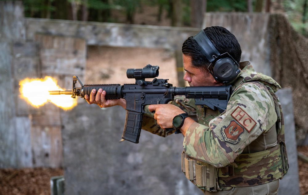 A military or law enforcement personnel is actively firing an AR-platform rifle at what appears to be an outdoor shooting range. A muzzle flash is clearly visible at the barrel, indicating the weapon is being discharged. The individual is wearing full tactical gear including OCP/MultiCam camouflage, a plate carrier, hearing protection, and eye protection.