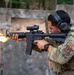 A military or law enforcement personnel is actively firing an AR-platform rifle at what appears to be an outdoor shooting range. A muzzle flash is clearly visible at the barrel, indicating the weapon is being discharged. The individual is wearing full tactical gear including OCP/MultiCam camouflage, a plate carrier, hearing protection, and eye protection.