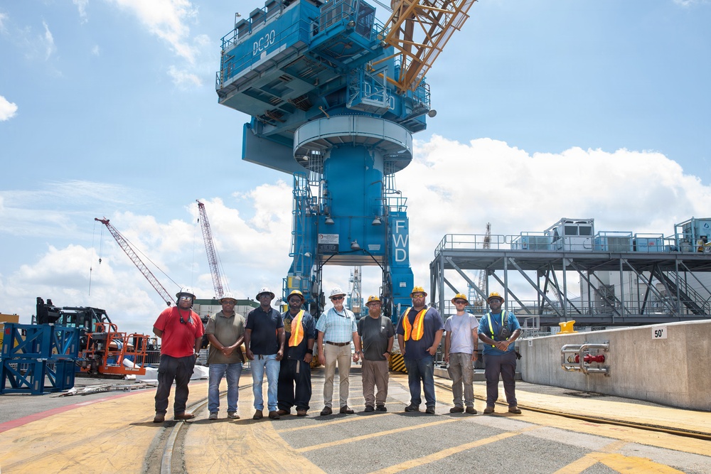 A group of approximately eleven workers and personnel pose for a group photo at an industrial port or shipyard facility. They are standing in front of a large blue harbor crane marked 'DC30' and 'FWD'. The setting appears to be an active heavy industrial worksite with multiple cranes and equipment visible.
