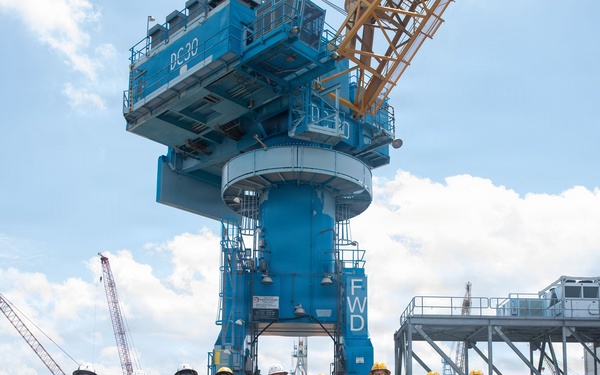A group of approximately eleven workers and personnel pose for a group photo at an industrial port or shipyard facility. They are standing in front of a large blue harbor crane marked 'DC30' and 'FWD'. The setting appears to be an active heavy industrial worksite with multiple cranes and equipment visible.