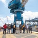 A group of approximately eleven workers and personnel pose for a group photo at an industrial port or shipyard facility. They are standing in front of a large blue harbor crane marked 'DC30' and 'FWD'. The setting appears to be an active heavy industrial worksite with multiple cranes and equipment visible.