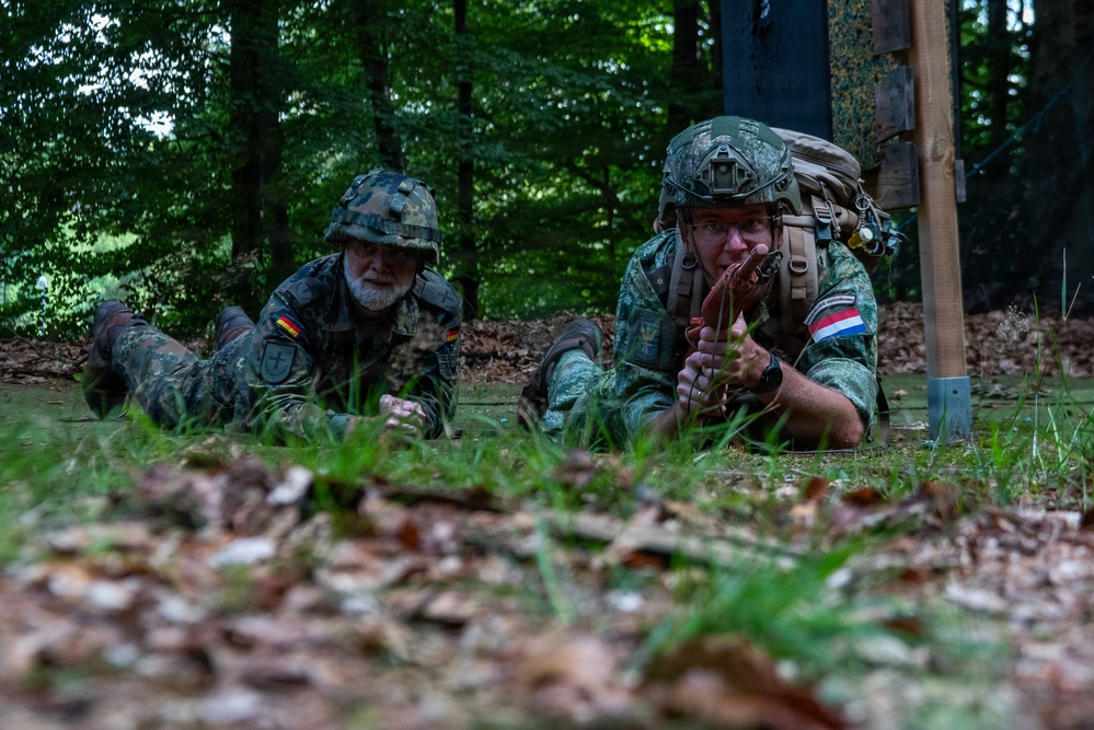 Two soldiers in camouflage uniforms are lying prone on the ground in a forested area, appearing to be in a tactical training exercise. The soldier on the right wears a Netherlands flag patch and is holding what appears to be a red training device, while the soldier on the left wears a German flag patch. Both are wearing helmets and military gear consistent with a joint NATO field exercise.