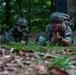 Two soldiers in camouflage uniforms are lying prone on the ground in a forested area, appearing to be in a tactical training exercise. The soldier on the right wears a Netherlands flag patch and is holding what appears to be a red training device, while the soldier on the left wears a German flag patch. Both are wearing helmets and military gear consistent with a joint NATO field exercise.