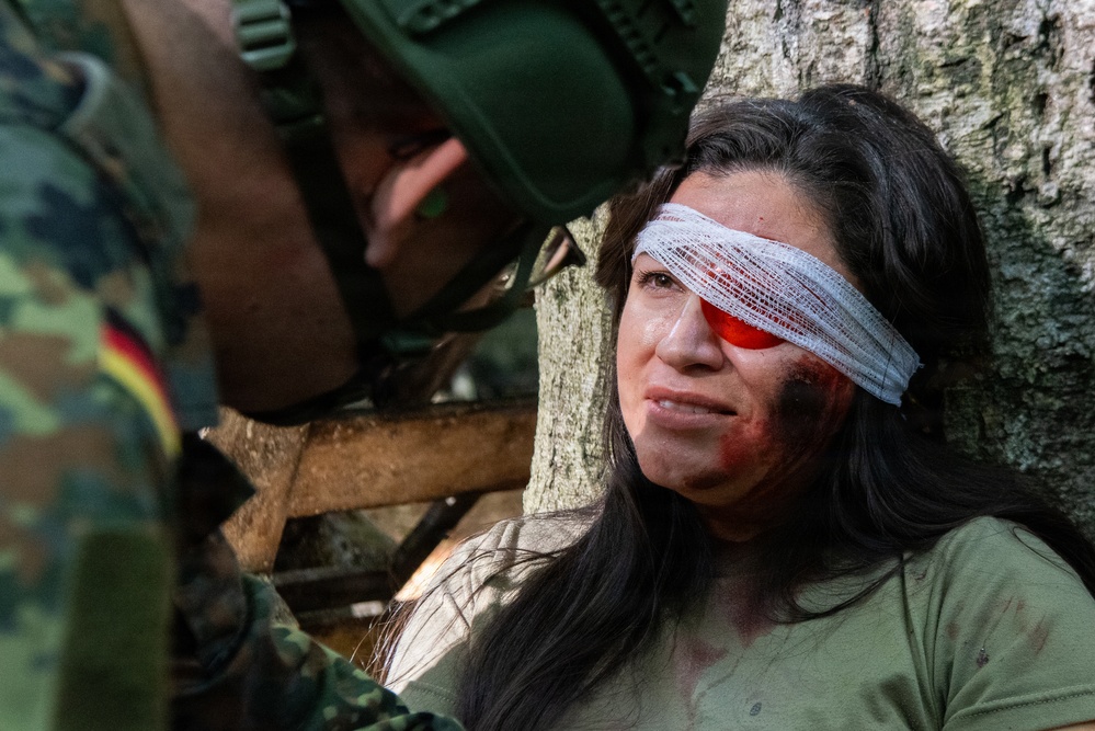 A military medical training or exercise scenario showing a woman in an olive green shirt with simulated facial injuries being attended to by a soldier wearing German Bundeswehr camouflage uniform. The woman has a white bandage wrapped around her head covering one eye, with a red eye shield visible underneath, along with simulated blood and bruising on her face. This appears to be a combat casualty care drill or MEDEVAC training exercise.