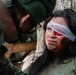 A military medical training or exercise scenario showing a woman in an olive green shirt with simulated facial injuries being attended to by a soldier wearing German Bundeswehr camouflage uniform. The woman has a white bandage wrapped around her head covering one eye, with a red eye shield visible underneath, along with simulated blood and bruising on her face. This appears to be a combat casualty care drill or MEDEVAC training exercise.