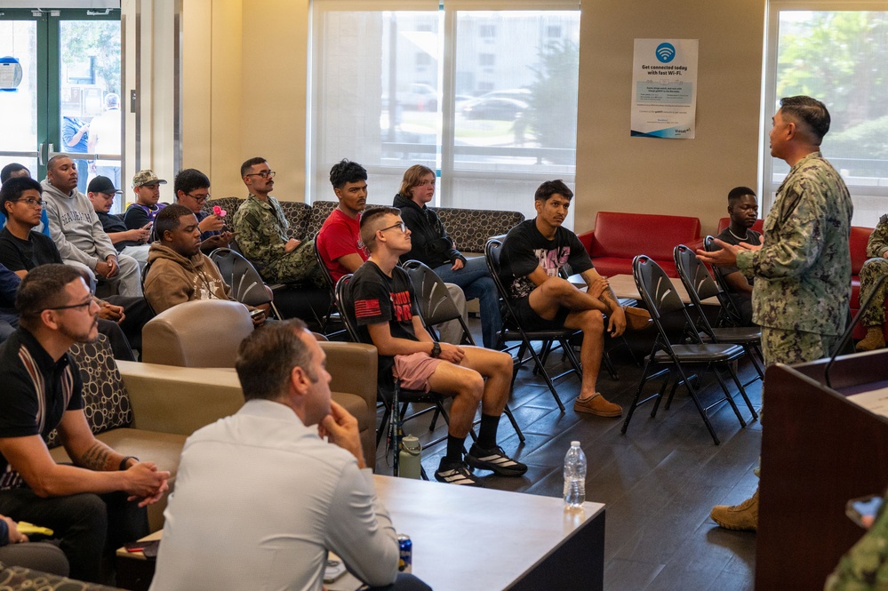 A person in U.S. Navy camouflage uniform (NWU) is addressing a group of approximately 20 individuals seated in folding and lounge chairs in what appears to be a waiting room or common area. The audience consists of a diverse group of young civilians and at least one other service member, suggesting this may be a military recruitment briefing or orientation session. A Wi-Fi advertisement sign is visible on the wall in the background.