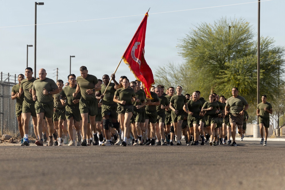 A large formation of military personnel in olive green PT (physical training) uniforms are conducting a group run on a paved road. One individual in the center carries a red unit guidon flag, consistent with U.S. Marine Corps formations. The setting appears to be a military installation in an arid/desert environment.