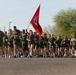 A large formation of military personnel in olive green PT (physical training) uniforms are conducting a group run on a paved road. One individual in the center carries a red unit guidon flag, consistent with U.S. Marine Corps formations. The setting appears to be a military installation in an arid/desert environment.