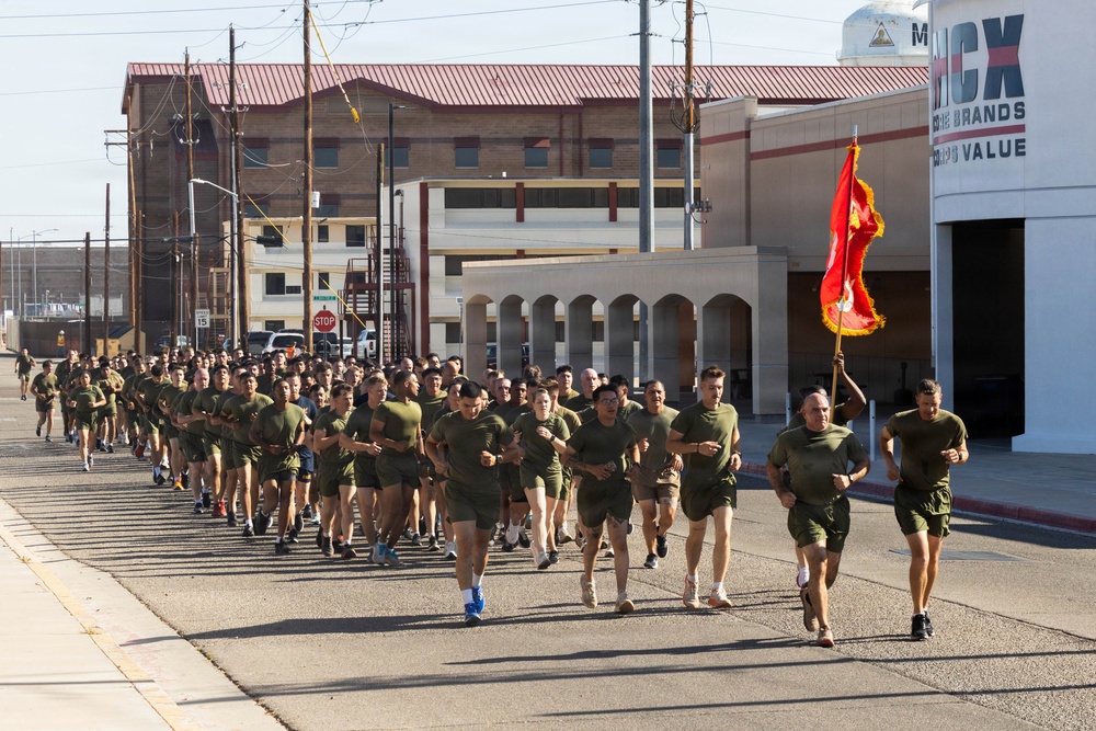 A large formation of military personnel, dressed in olive green physical training uniforms, are conducting a unit run along a road on what appears to be a military installation. A guidon bearer at the front right carries a red and gold flag consistent with U.S. Marine Corps colors. The background shows base infrastructure including a building with 'MCX BRANDS VALUE' signage, indicating a Marine Corps Exchange facility.
