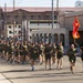 A large formation of military personnel, dressed in olive green physical training uniforms, are conducting a unit run along a road on what appears to be a military installation. A guidon bearer at the front right carries a red and gold flag consistent with U.S. Marine Corps colors. The background shows base infrastructure including a building with 'MCX BRANDS VALUE' signage, indicating a Marine Corps Exchange facility.