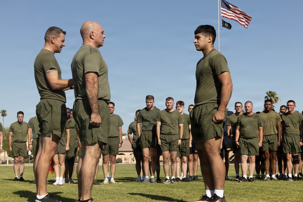 A group of military personnel in olive drab physical training uniforms (green t-shirts and shorts) are assembled on a grass field. Two senior individuals appear to be conducting an inspection or award ceremony with a third individual standing at attention. An American flag and what appears to be a POW/MIA flag are visible on a flagpole in the background.