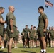 A group of military personnel in olive drab physical training uniforms (green t-shirts and shorts) are assembled on a grass field. Two senior individuals appear to be conducting an inspection or award ceremony with a third individual standing at attention. An American flag and what appears to be a POW/MIA flag are visible on a flagpole in the background.