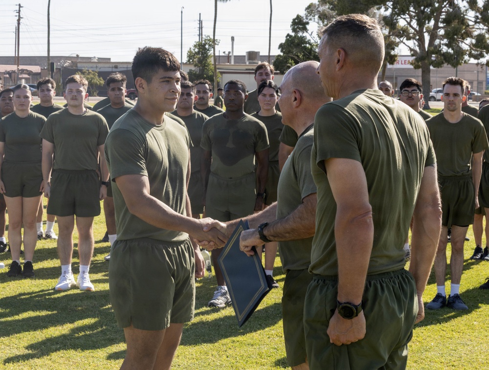 A military ceremony is taking place outdoors on a grass field, where a young service member in olive drab physical training (PT) gear is shaking hands with a senior military official who is holding a framed certificate or award. A large formation of similarly uniformed personnel stands at attention in the background. The setting appears to be a U.S. military installation, likely a Marine Corps base based on the olive green PT uniforms.
