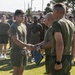 A military ceremony is taking place outdoors on a grass field, where a young service member in olive drab physical training (PT) gear is shaking hands with a senior military official who is holding a framed certificate or award. A large formation of similarly uniformed personnel stands at attention in the background. The setting appears to be a U.S. military installation, likely a Marine Corps base based on the olive green PT uniforms.