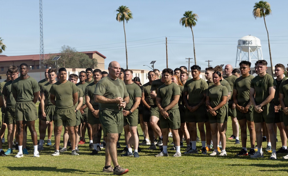A group of military personnel in olive green physical training (PT) uniforms are assembled on a grass field at what appears to be Marine Corps Air Station (MCAS), as indicated by the water tower in the background. A senior individual, likely an officer or senior NCO, stands facing the group and appears to be addressing them. The setting suggests a physical training formation or briefing event.