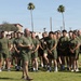 A group of military personnel in olive green physical training (PT) uniforms are assembled on a grass field at what appears to be Marine Corps Air Station (MCAS), as indicated by the water tower in the background. A senior individual, likely an officer or senior NCO, stands facing the group and appears to be addressing them. The setting suggests a physical training formation or briefing event.