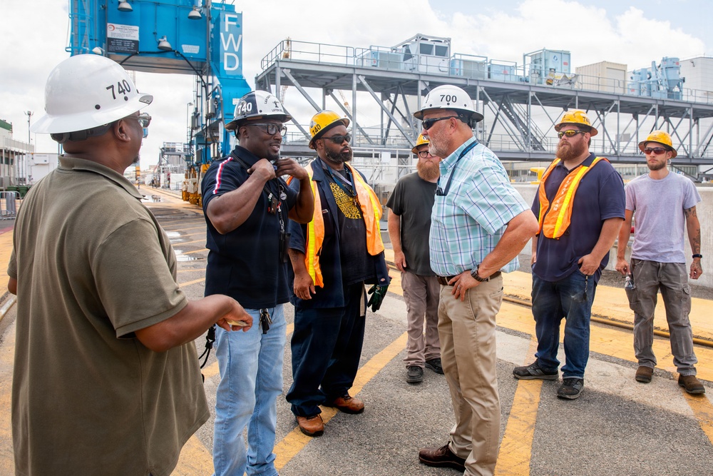 A group of approximately seven workers are gathered at an industrial port or shipyard facility, engaged in discussion. Several individuals are wearing hard hats with numbers '740' and '700' visible, along with orange high-visibility safety vests. The setting includes heavy industrial infrastructure including a blue crane structure marked 'FWD' in the background.