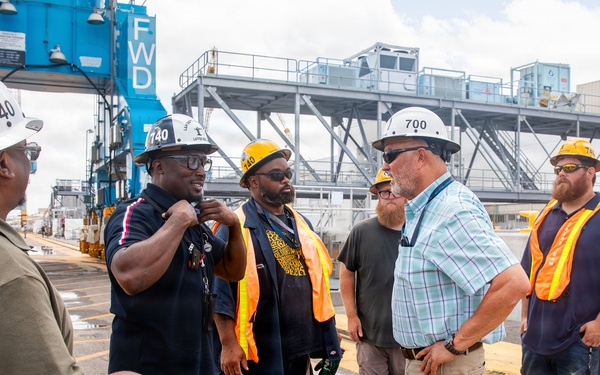 A group of approximately seven workers are gathered at an industrial port or shipyard facility, engaged in discussion. Several individuals are wearing hard hats with numbers '740' and '700' visible, along with orange high-visibility safety vests. The setting includes heavy industrial infrastructure including a blue crane structure marked 'FWD' in the background.