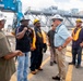 A group of approximately seven workers are gathered at an industrial port or shipyard facility, engaged in discussion. Several individuals are wearing hard hats with numbers '740' and '700' visible, along with orange high-visibility safety vests. The setting includes heavy industrial infrastructure including a blue crane structure marked 'FWD' in the background.