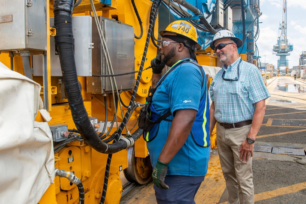 Two workers are standing beside large yellow industrial machinery at what appears to be a port or construction site. One worker wearing a yellow hard hat and blue safety vest is speaking on a corded phone attached to the equipment, while a second worker in a white hard hat and plaid shirt looks on smiling. A crane and port infrastructure are visible in the background.